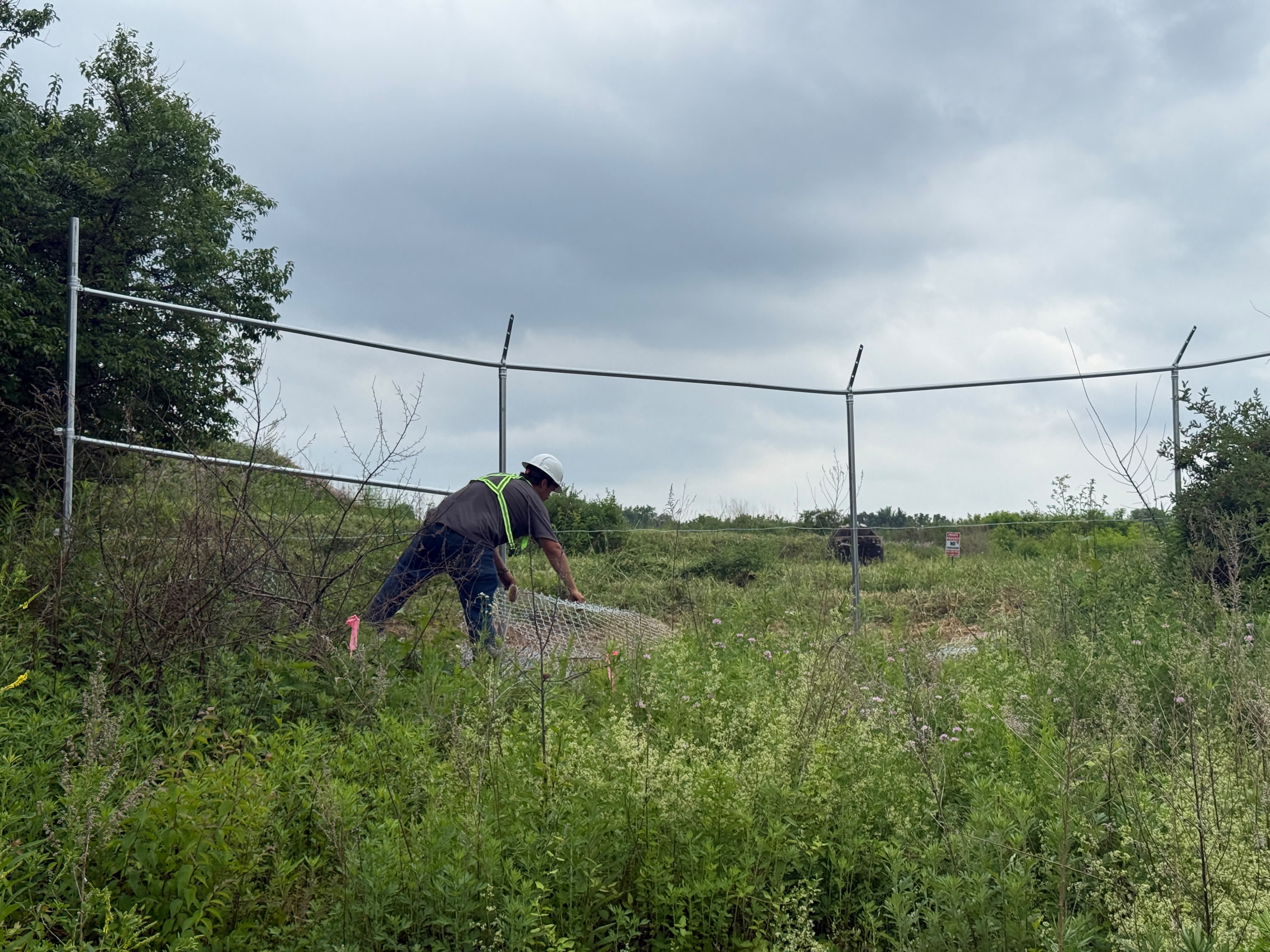 A construction worker in a hard hat installing chain-link fencing at the site, without visible additional PPE.